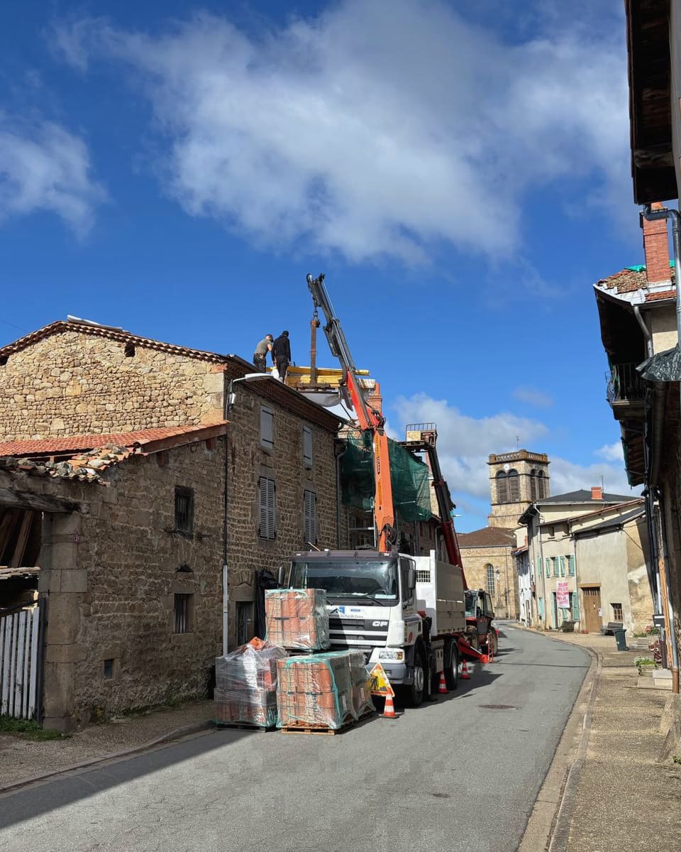 Chantier de couverture dans une rue de bourg : camion-grue, palettes de tuiles au sol et maison en pierre en cours de réfection.